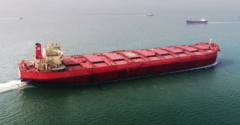 An aerial view of a massive red Pan Ocean dry bulk carrier vessel sailing on the ocean. Represents the company's core shipping fleet and raw material transport capabilities.